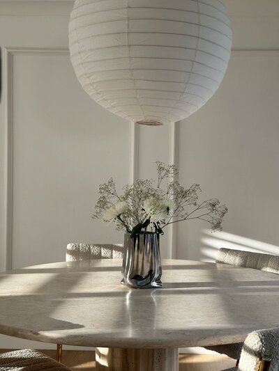Round cream natural stone dining table surrounded by white bouclé dining chairs. A large Noguchi-style paper pendant hangs above, while a silver organic-shaped vase with white flowers and baby's breath completes the setting.