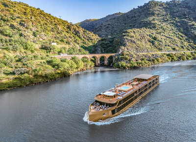 A modern river cruise ship sails along a winding river between green hills, passing near an old stone bridge.