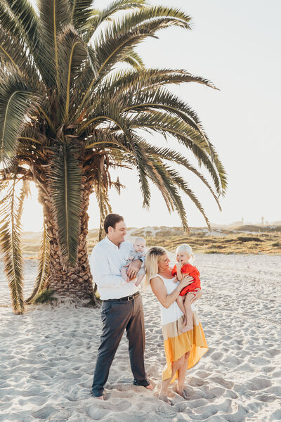 A family of four laughs together while at their beach photography session with a giant palm tree in the background