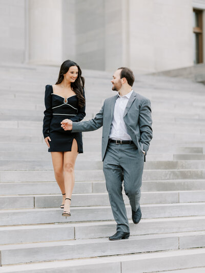 couple walks down steps at Arkansas State Capitol in Little Rock, Arkansas wearing grey suit and short black dress