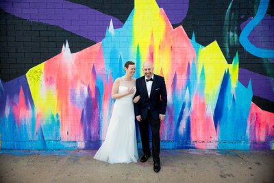 A wedding couple standing arm in arm in front of a colorful mural.