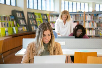 Girl in a olive shirt and beige jacket studying for her PA exams in a library with two other women in the background