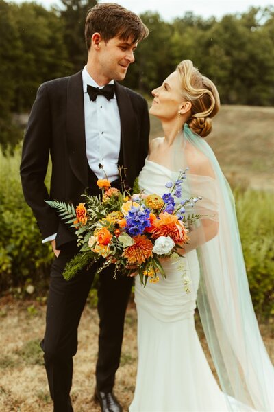 Chic bride and groom look into each others' eyes while standing in front of woods. Groom in bowtie, bride with long elegant train, holding large bold floral bouquet.
