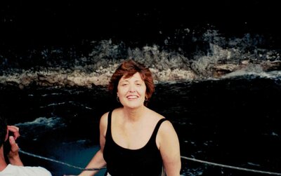 A smiling woman in a black swimsuit standing in the ocean with volcanic rocks in the background in Kauai, capturing a moment of joy and reflection during a meaningful mother-daughter trip.