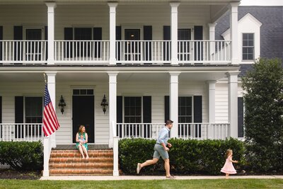 A father runs after his daughter as his wife sits on the porch with newborn baby.