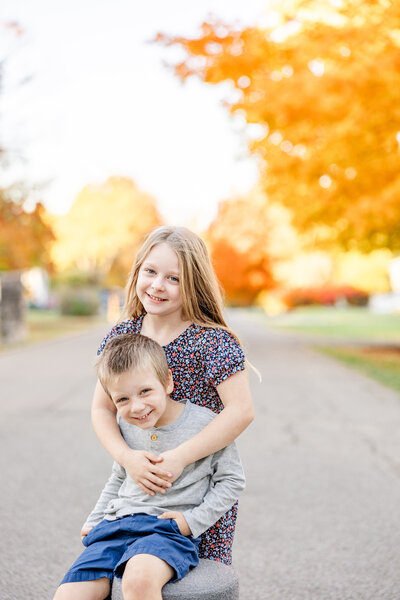 Brother sitting on a stool with his hands in his pockets in the street on an orange Fall evening with his sisters arms wrapped around him in a hug while they both smile at the camera.