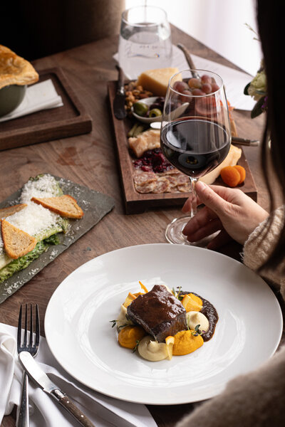 a woman enjoying a glass of wine while sitting at a table with a plate of elevated steak and vegetables