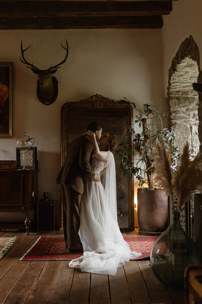 Cinematic fine art portrait of a bride seated in soft window light with her groom leaning in for a kiss inside a historic Scottish castle, photographed by wedding photographer Aly Robinson.