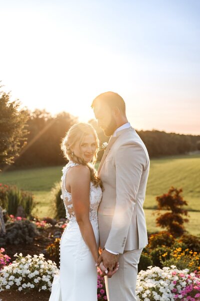 A bride and groom stand hand in hand during golden hour, with the warm setting sun glowing behind them in a landscaped garden filled with white and pink flowers. The bride’s fitted lace gown features a sheer back and floral appliqué, while the groom wears a light beige suit and looks down tenderly at her as she smiles softly toward the camera.