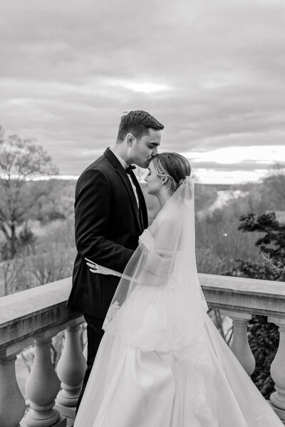 Wedding at the Estate at River Run in Maidens, VA | Wedding Photographer in Richmond | black and white images of bride with flowing veil and groom in a warm embrace on the balcony of the Estate at River Run as the groom gently kisses the bride on her forehead.