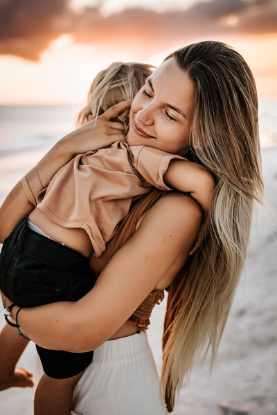 Family Photographer, a mother holds her baby boy near the ocean at sunset