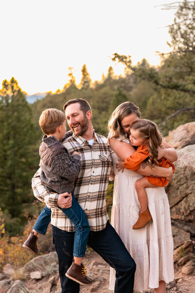 Mom and dad each hold a kid and snuggle them on front of evergreens for their family photos.