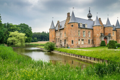 A historic brick castle with pointed towers stands beside a tranquil moat, surrounded by lush greenery under a cloudy sky—a sight that any European travel specialist would recommend for an authentic cultural journey.