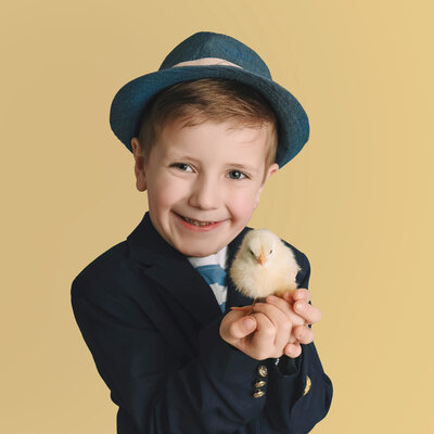 A boy with a hat and suit jacket holds a baby chick and smiles