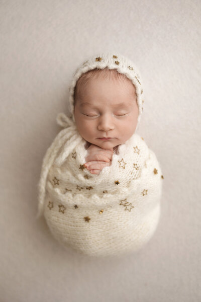 Newborn baby swaddled in a white knitted wrap adorned with golden stars, wearing a matching white knitted bonnet. The baby is peacefully sleeping on a white blanket.