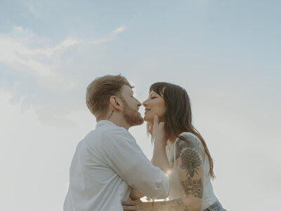 a close up photo of a couple, where the man is holding his girlfriends chin and pulling her in close for a kiss. the backdrop of the beautiful blue sky at Woodlawn State Park outside of Buffalo, NY. Photo by Dash and Boheme Photography. 