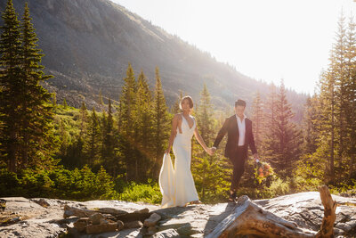 Shannon Lee, a Colorado elopement photographer capturing a couple walking hand in hand through a stunning mountain landscape, with the sun shining brightly behind them.
