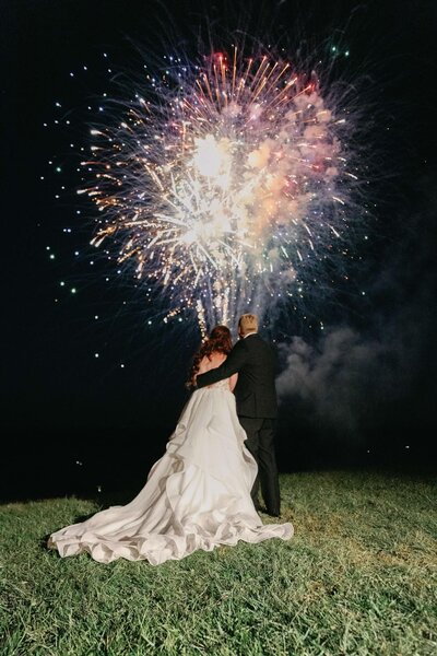 Bride and groom looking at fireworks standing on a hill with the picture being taking behind the couple with the fireworks going off. 