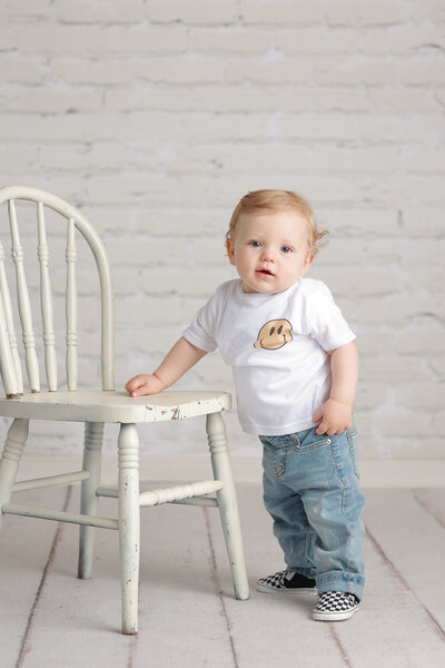 Lehigh Valley First Birthday Photographer one year old boy portrait from his first birthday cake smash standing at a white chair, wearing a white tshirt with a yellow happy face on it and jeans with checkered Vans shoes and a backdrop of white brick and white wood floor under him