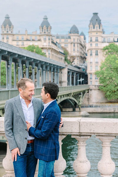 Gay couple embracing and laughing together near the Eiffel Tower at Bir-Hakeim Bridge during a romantic Paris couples photoshoot.