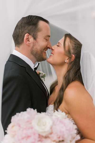 A classic photo of a bride and groom in true to life colors. The bride and groom are rubbing noses and laughing while the veil is over their heads. 
