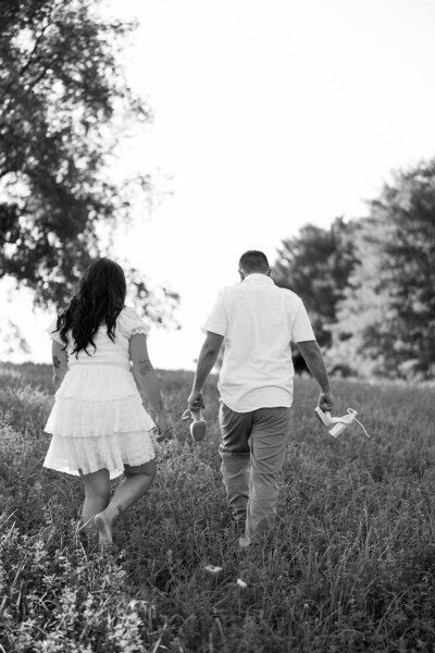 A couple walking away from the camera in an open field during their Detroit engagement session, with the groom holding her shoes while she walks barefoot.