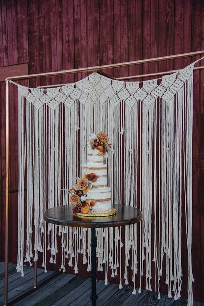 Photo shows a three tier wedding cake with a naked style frosting (some cake is showing through). The cake is decorated with fresh flowers in shades of fall colors, and various candied fruits. There's a smaller cluster on the top right of the cake, and a larger cascade on the bottom left side.