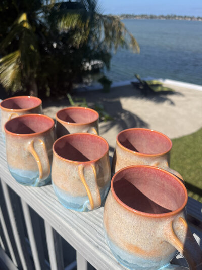 A group of mugs sit on a table, they are red on the inside and orange, cream, blue and turquoise on the outside