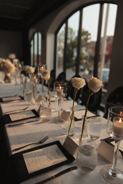 wedding table details with white roses in bud rose vases, flay lay stationary menus with candle lit dinner on family style long table for wedding reception