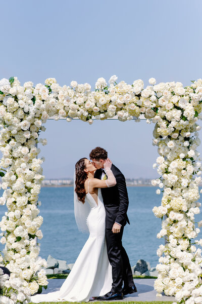 Bride and groom kiss at their wedding reception surrounded by lush white floral arch with Pacific Ocean behind them