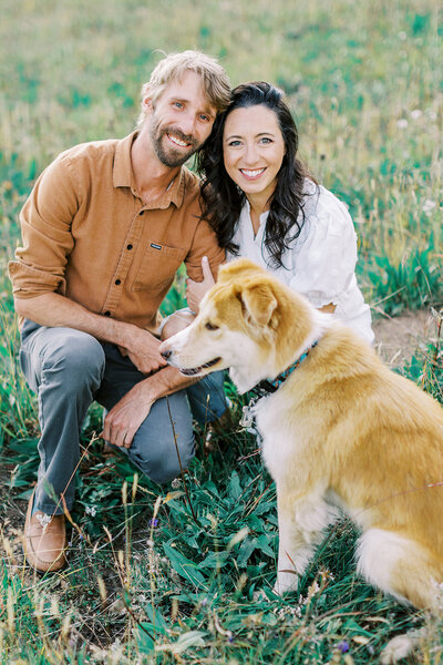 A happy couple in white and brown snuggle in the grass with their brown dog sitting in front of them