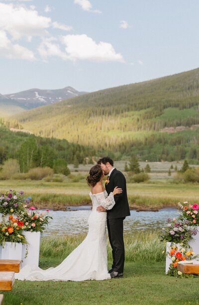 Bride and groom sharing a kiss by the lake at Camp Hale in Vail, Colorado, surrounded by mountain views and vibrant florals.