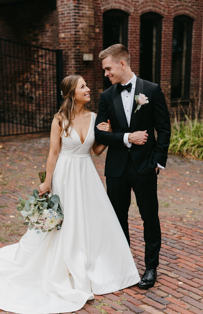Bride and groom with their arms interlocked smiling at each other in front of a brick wall