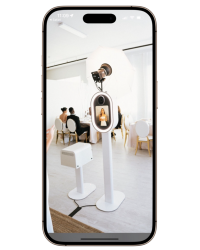 A sleek digital photo booth on display at a wedding reception, showing a live preview of a guest posing, with elegant white chairs and tables in the background.
