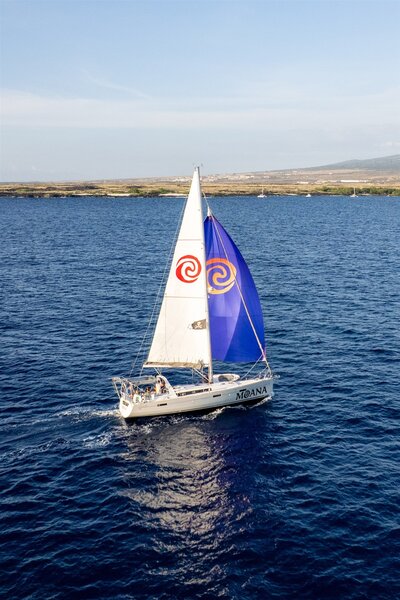 Aerial view of the Moana sailboat cruising over deep blue water off the Big Island of Hawaii, with colorful sails and a clear coastline in the background.