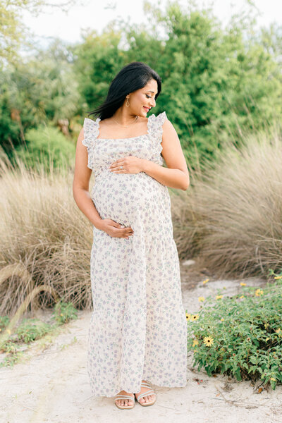 Pregnant mother wearing floral dress holding baby bump and smiling down at shoulder outside in Tampa, Florida