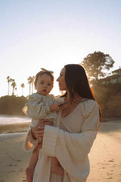 Mother holding her baby at golden hour on the beach in Laguna Beach, Orange County. Candid lifestyle family photo by Orange County photographer Maria Alcantara.