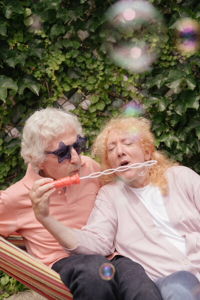 Two older partners stand close together, blowing bubbles from the same bubble wand. One partner wears blue star-shaped glasses. An ivy-covered fence is visible behind them.