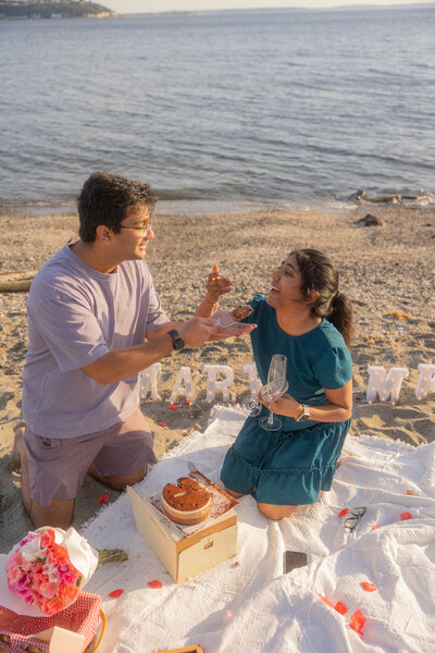 Man feeding woman cake after a heartfelt proposal at the beach in front of a Marry me sign