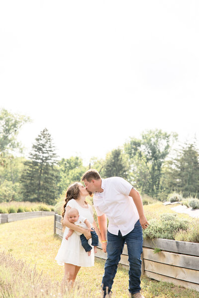 Mom and dad kiss, holding hands while mom holds baby boy in a lavender field in Miamisburg, Ohio.