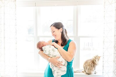 woman stand by window holding baby during in hone newborn photo session with Sara Sniderman  Photography in Natick Massachusetts