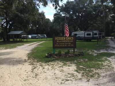 A campground entrance sign reads "Hidden Oaks RV and Camp Sites LLC" with contact information, ideal for those seeking Mayo Florida camping. An American flag waves proudly beside it. RVs rest under large trees, with a gravel path inviting guests to explore the serene surroundings.
