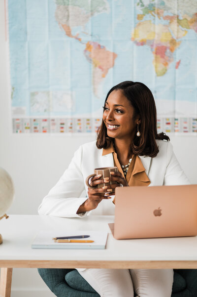 A woman in a white blazer and brown pants sits at a desk with a laptop and globe, planning destination weddings.