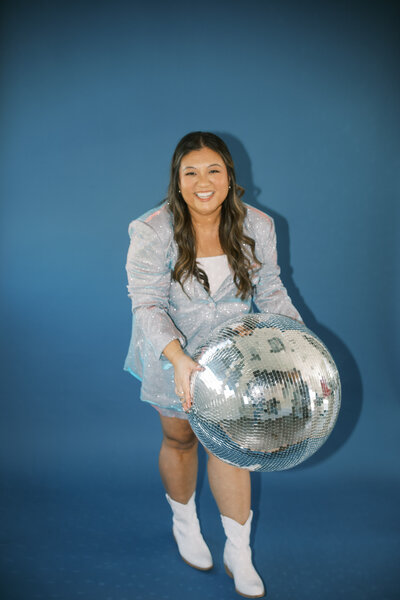 Monique Anne in a sparkly blazer and white cowgirl boots holding a disco ball in front of a blue background