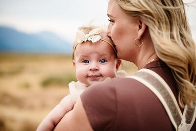 A mother kissing her baby on the forehead while baby blows spit bubbles. 