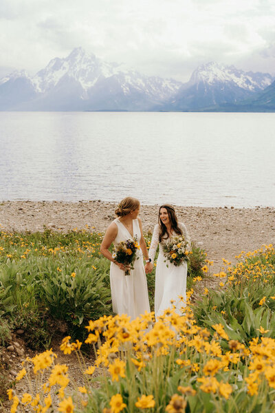 two brides holding hands while walking next to a lake with mountains in the background