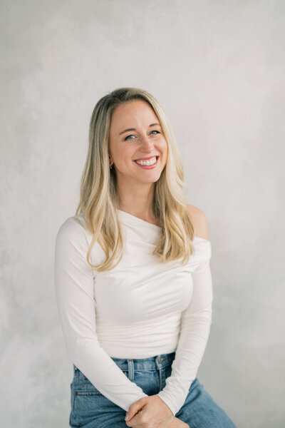 smiling photographer, wearing jeans and an off-shoulder white Abercrombie shirt, sitting casually for a portrait