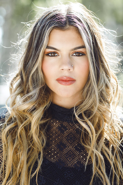 Strong and striking senior headshot featuring a young woman with caramel blonde hair, bold makeup, and a black lace top. Sunlight adds contrast for a fierce, confident expression.