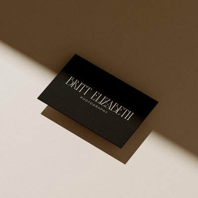 A black business card with the text "BRIT ELIZABETH PHOTOGRAPHY," created by a talented branding designer, is placed on a surface partially illuminated by light.