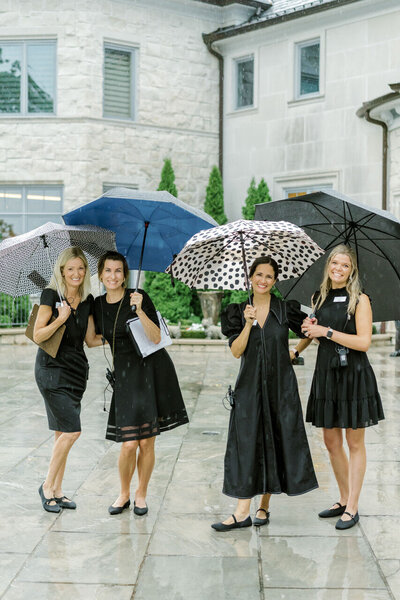 Four women dressed in black holding umbrellas and smiling.